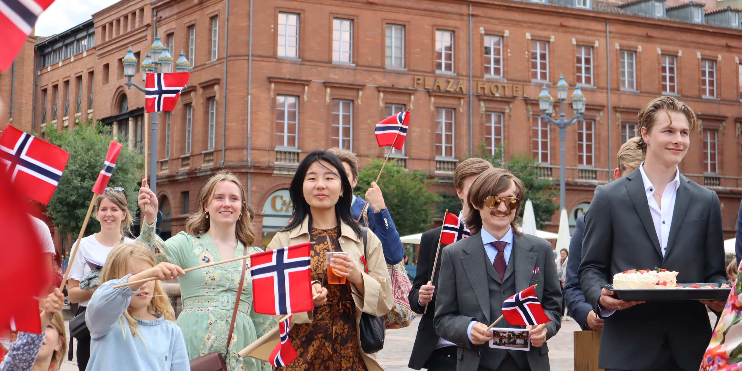 Norske studenter i finstasen med flagg og kake på 17. mai, foran teglsteinsfasadene i Toulouse